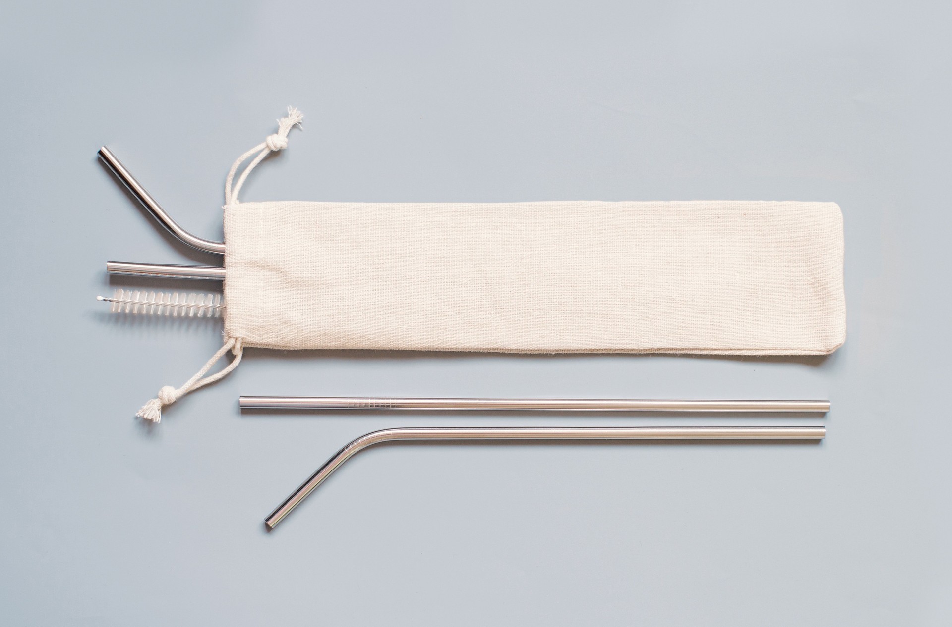 Reusable stainless steel straws and cleaning brush with cream cotton bag on grey background.
